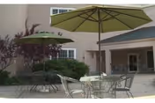 Outdoor patio area with metal tables and chairs under large green umbrellas, surrounded by bushes and a building with windows and a covered entrance.
