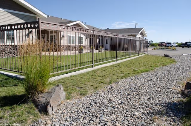 Exterior view of a single-story assisted living facility building with beige siding and stone accents. A metal fence runs along a grassy area with some ornamental grasses and rocks. A gravel pathway is visible in the foreground, and several parked cars are seen in the background under a clear blue sky.