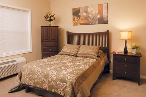 A cozy bedroom with a double bed covered in a floral patterned bedspread and matching pillows. There is a wooden headboard, a nightstand with a lamp and a small plant, a tall dresser with a flower vase on top, a window with blinds, and a wall painting above the bed.
