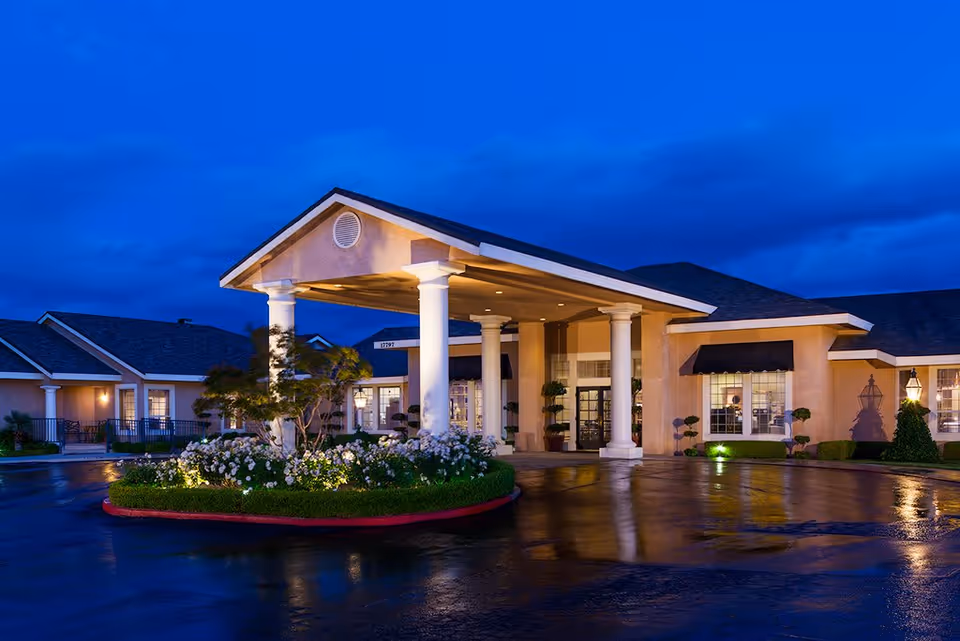 Exterior view of Sterling Commons Memory Care Community building at dusk with illuminated entrance featuring white columns, landscaped flower beds, and a wet driveway reflecting the lights.