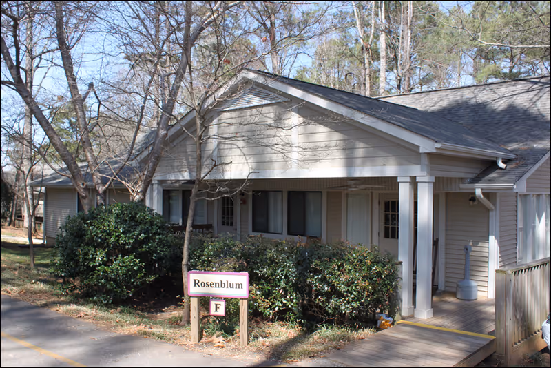 Single-story beige building with a covered front porch, shrubs and trees, and a small sign in front reading "Rosenblum" with the letter "F".