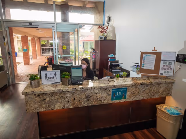 Reception area of Magnolia Post Acute Care with a woman wearing a pink face mask sitting behind a granite countertop desk. The desk has two small potted plants, a sign about social distancing, and a computer monitor. Behind the desk is a wooden cabinet with office supplies. The entrance door is glass, showing an outdoor patio area with brick columns and greenery.