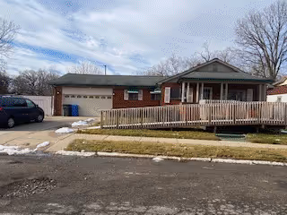 Single-story brick residential building with a wheelchair accessible wooden ramp leading to the front porch. There is a driveway with a parked car and three blue trash bins near the garage. The lawn is partially covered with patches of snow, and leafless trees are visible in the background under a cloudy sky.