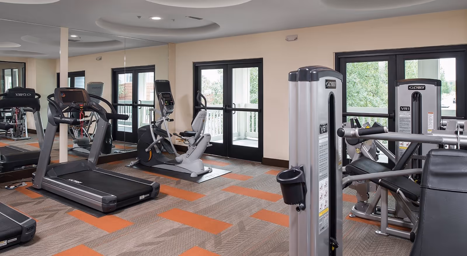 A fitness room with exercise equipment including treadmills, a recumbent bike, and weight machines. The room has large windows and glass doors allowing natural light to enter, with a patterned carpet featuring orange and beige tones.