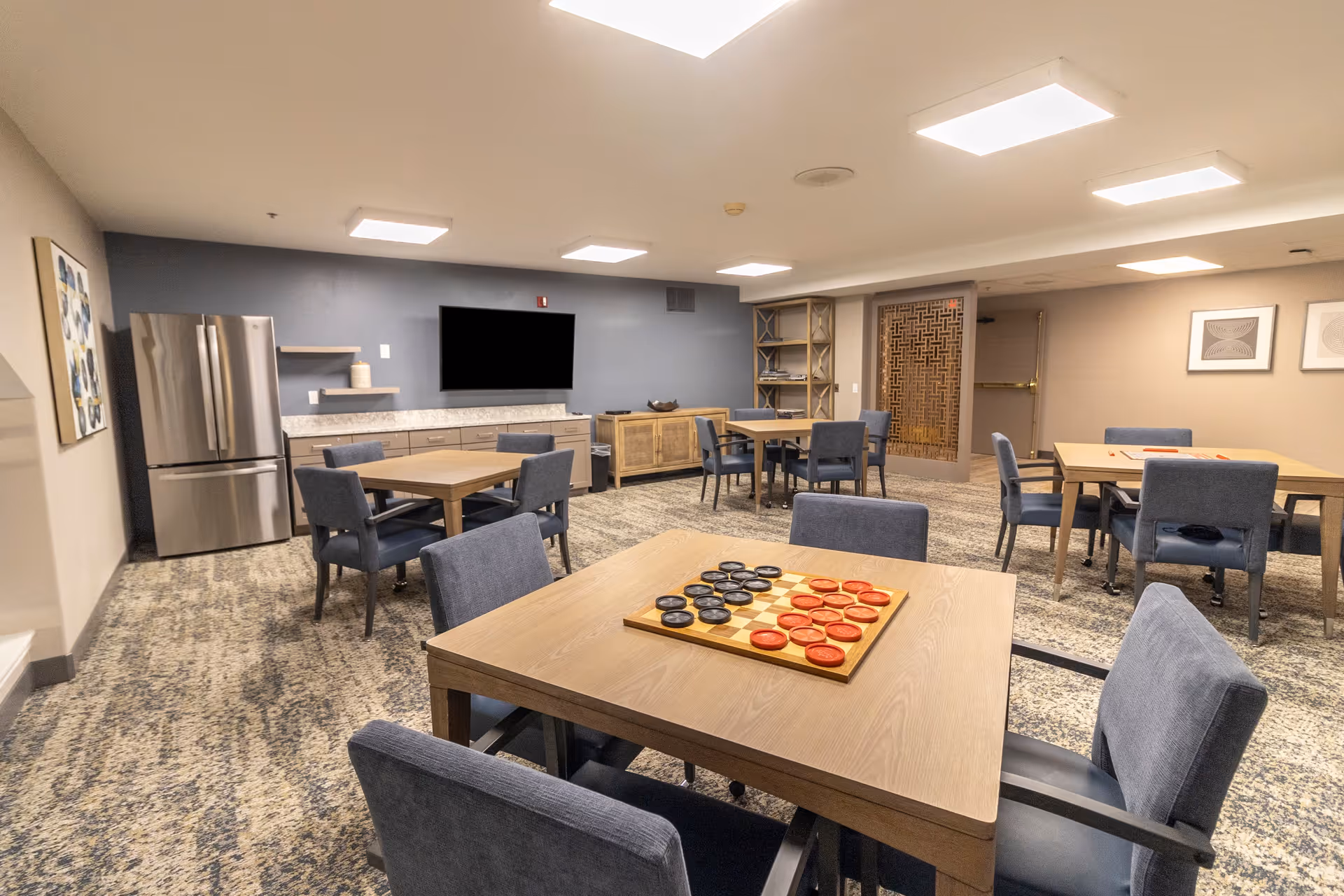 A well-lit common room with several wooden tables and blue upholstered chairs arranged around the room. One table in the foreground has a checkers game set up. The room features a large flat-screen TV mounted on a blue accent wall, a stainless steel refrigerator, and wooden cabinets. The carpet has a patterned design, and there are decorative framed artworks on the walls.