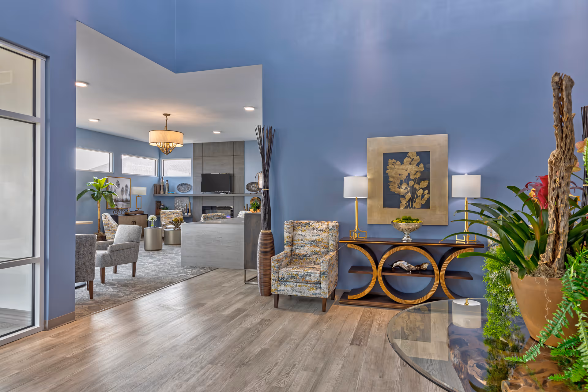 Bright communal living room with blue walls, seating areas, a decorative console table with lamps, and plants.