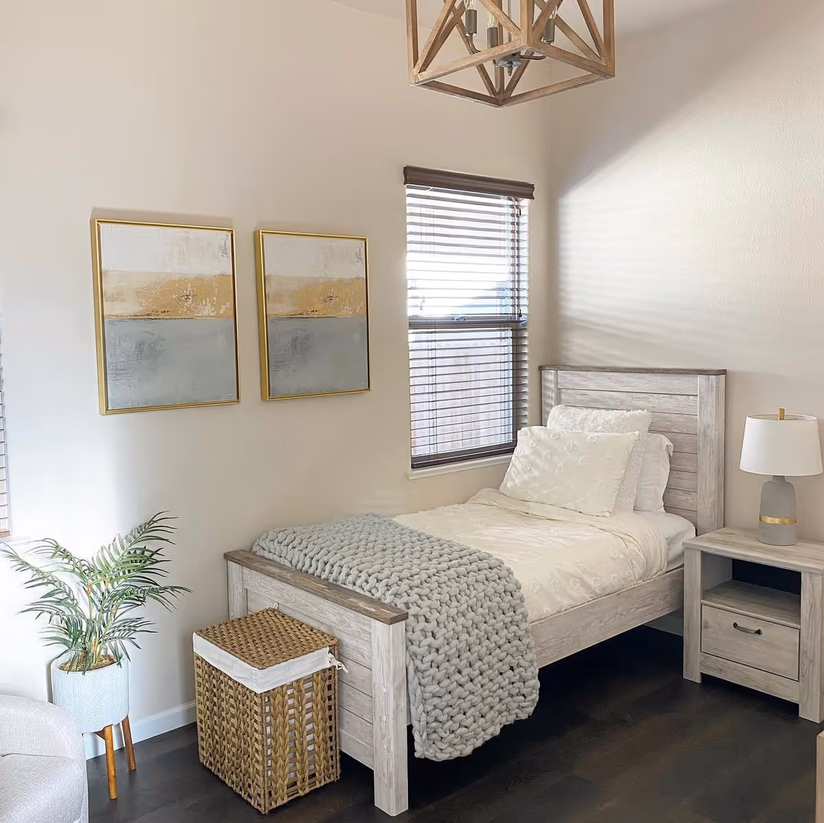 Bright, neatly decorated bedroom with a single bed, nightstand and lamp, framed wall art, window blinds, and a woven basket.