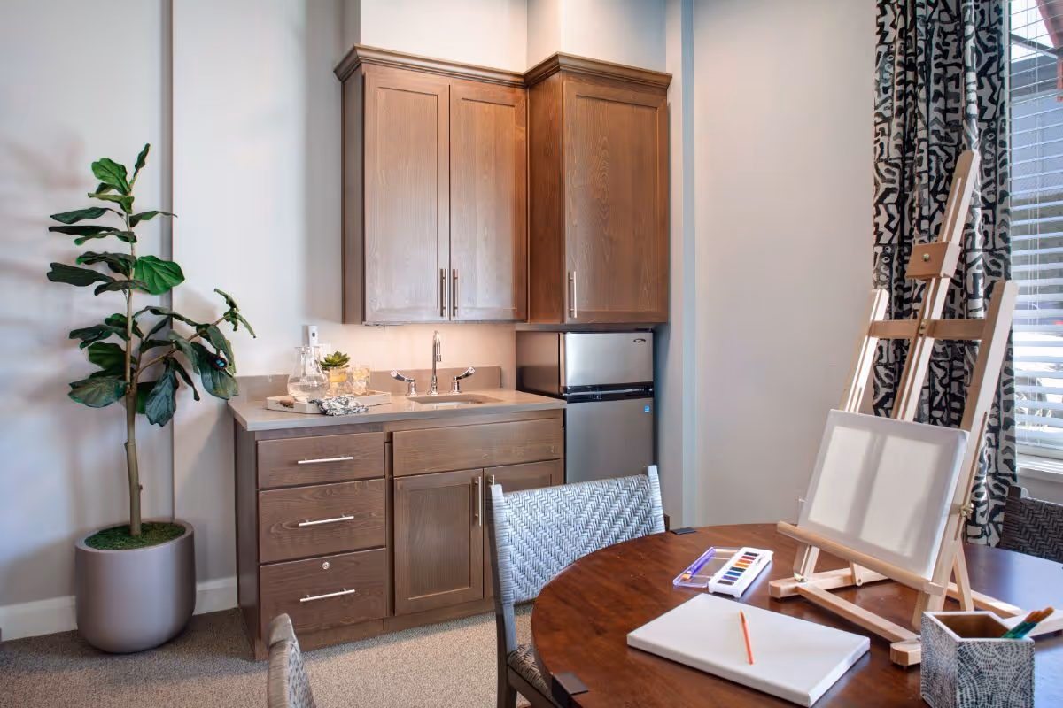 A small kitchenette area with wooden cabinets, a sink, and a mini refrigerator. Next to it is a round wooden table with an easel holding a blank canvas, watercolor paints, a notebook, and a pencil. A potted plant is placed to the left, and a window with patterned curtains is on the right.