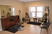 Interior view of a salon area in a senior living facility with a styling chair in front of a mirror and counter, two decorative chairs with patterned cushions near a window, a small table between the chairs, and additional seating along the wall.