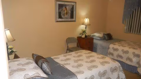 A bedroom with two twin beds covered in matching paisley-patterned bedspreads. Between the beds is a wooden nightstand with a lamp and a small plant. A folding chair is placed against the wall, and a framed picture hangs above the nightstand. The room has beige walls and a window with vertical blinds.