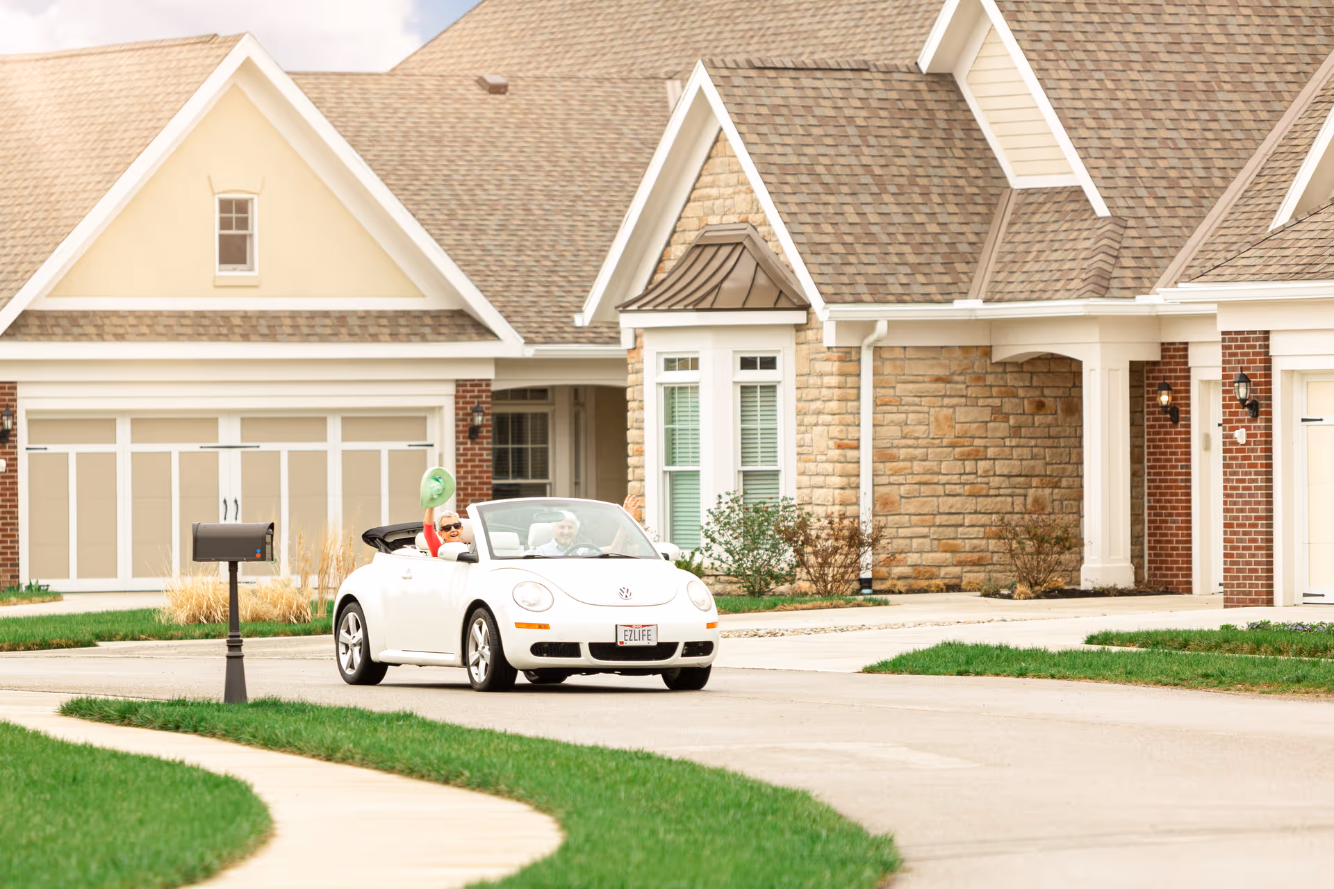 A white convertible with two occupants drives down a street in front of suburban homes with garages.