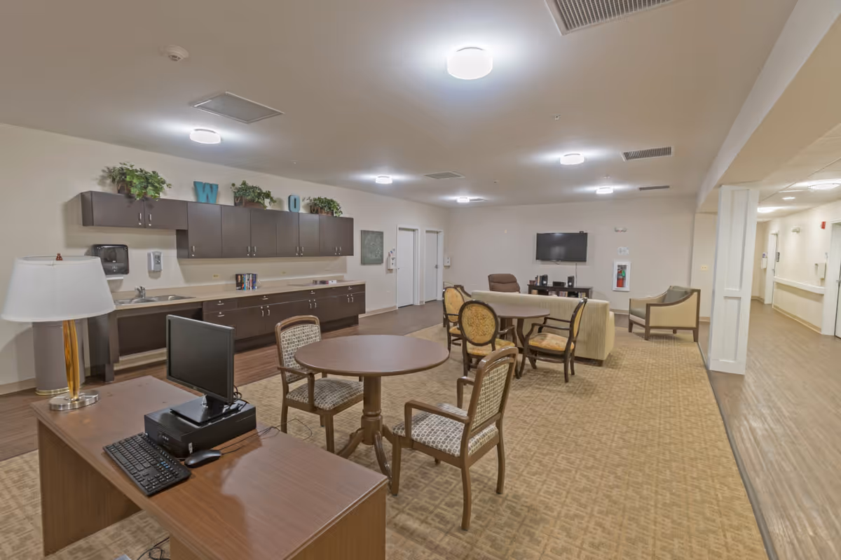 A spacious senior living common room with tables and chairs, a sofa and wall-mounted TV, kitchenette cabinets along the back wall, and a desk with a computer in the foreground.
