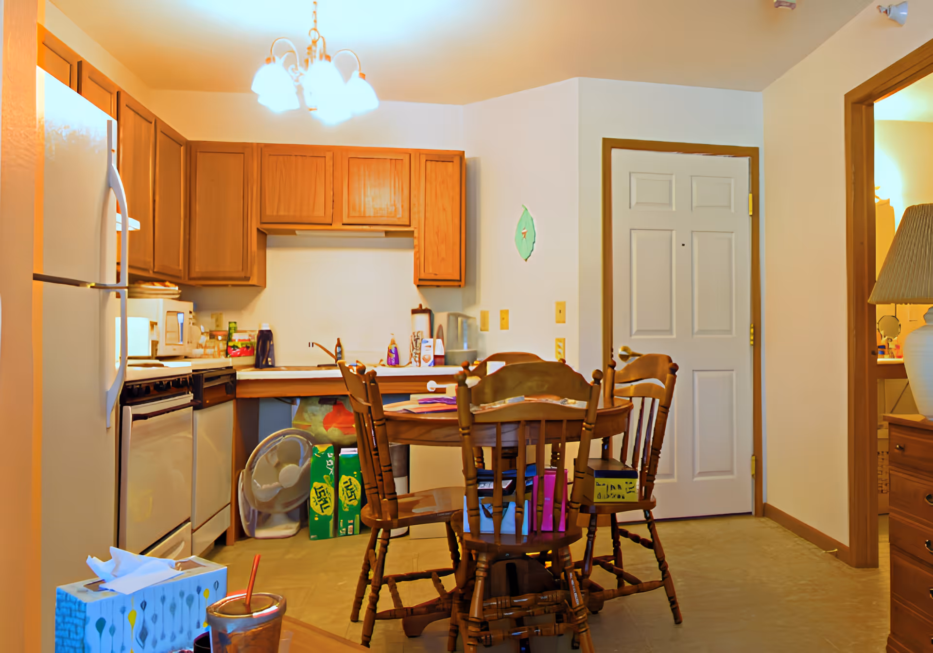 A small kitchen and dining area with wooden cabinets, a white refrigerator, stove, and dishwasher. A round wooden dining table with four wooden chairs is in the center. Various kitchen items and cleaning supplies are on the counter. A white door and an open doorway leading to another room are visible. A tissue box and a drink cup with a straw are on a nearby surface.