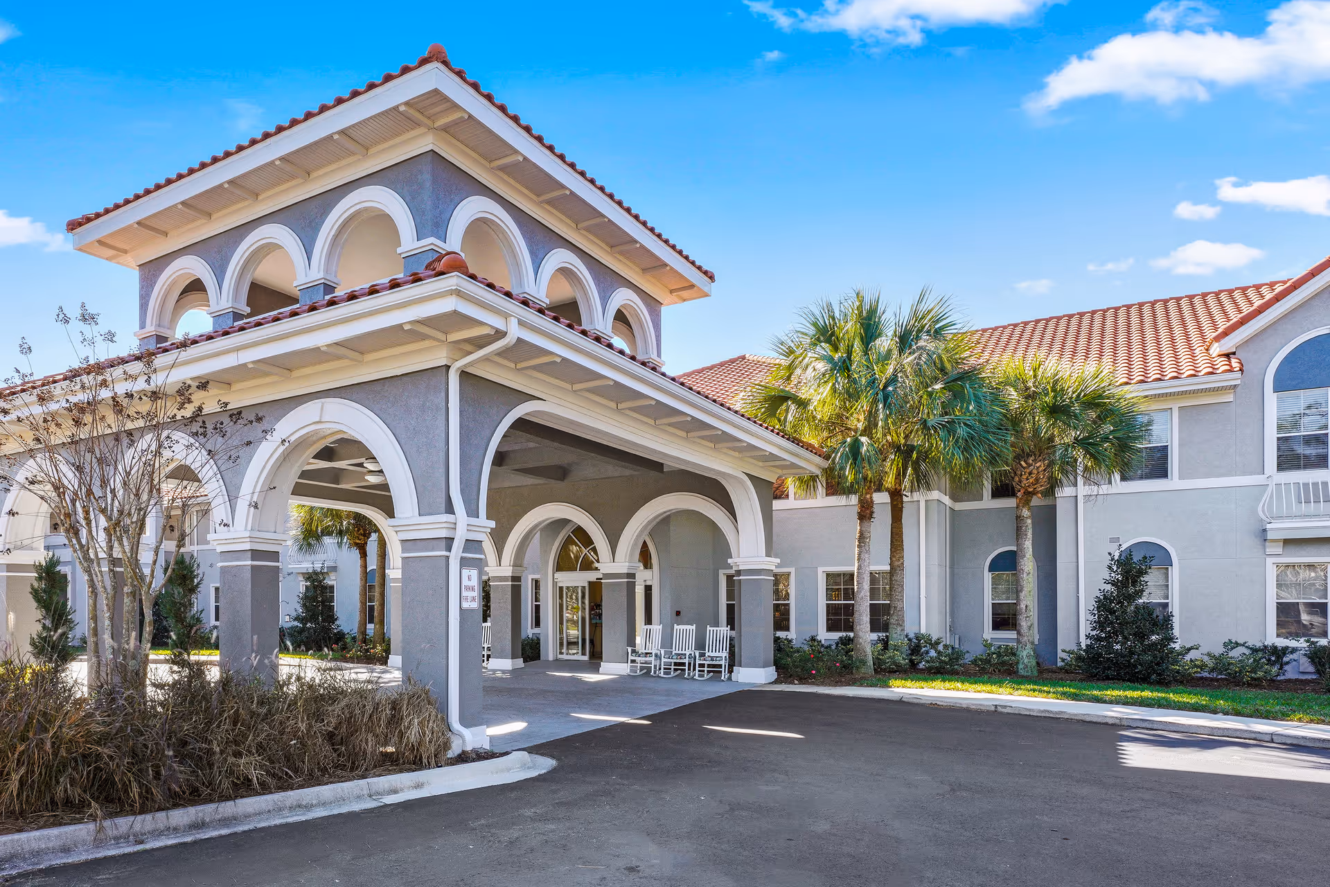 Exterior view of The Cove at Marsh Landing senior living facility showing a covered entrance with arches, rocking chairs, palm trees, and a clear blue sky.