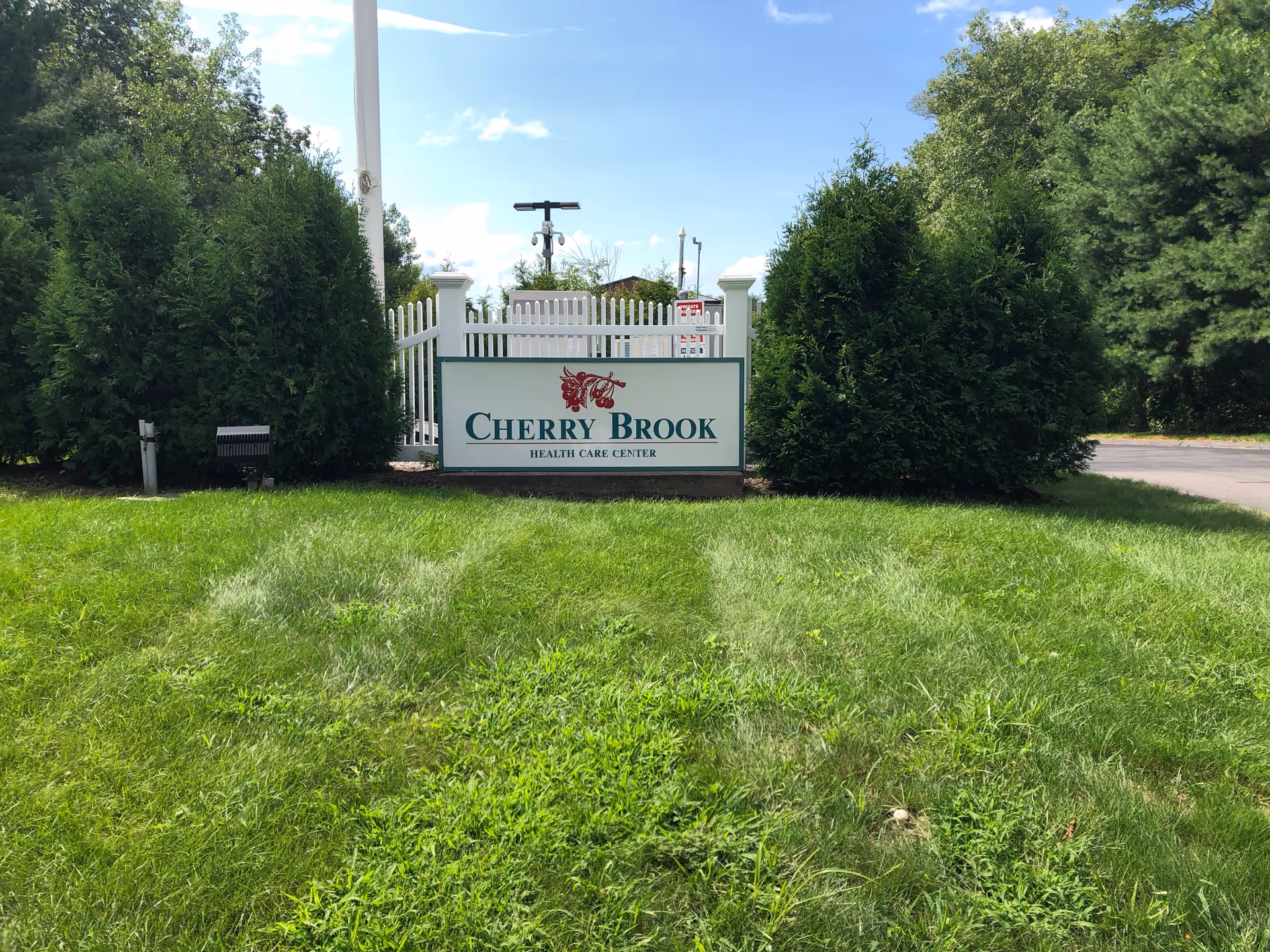 Outdoor view of a green grassy area with a white fence and a sign that reads 'Cherry Brook Health Care Center' surrounded by tall green bushes and trees under a partly cloudy blue sky.