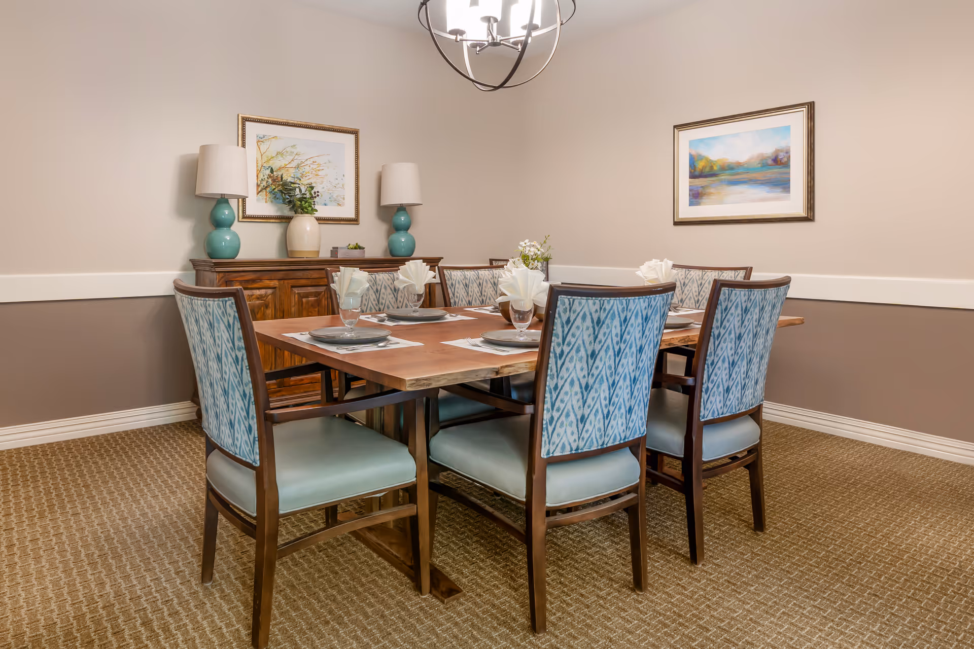 A dining room with a wooden table set for six, upholstered chairs, a sideboard with lamps, and framed artwork on the walls.