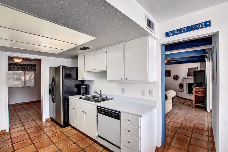 Kitchen with white cabinets, stainless steel refrigerator, double sink and dishwasher opening to a living room with a 'THIS WAY TO THE POOL' sign.