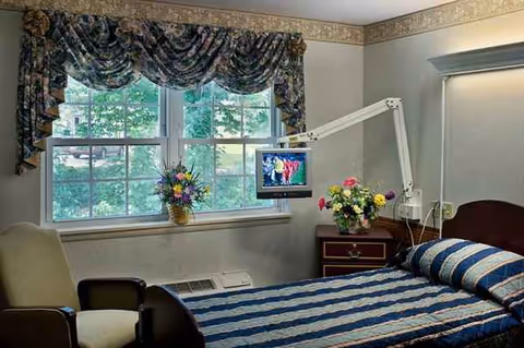 Patient bedroom in a nursing facility with an adjustable bed, monitor, flowers, and draped window.