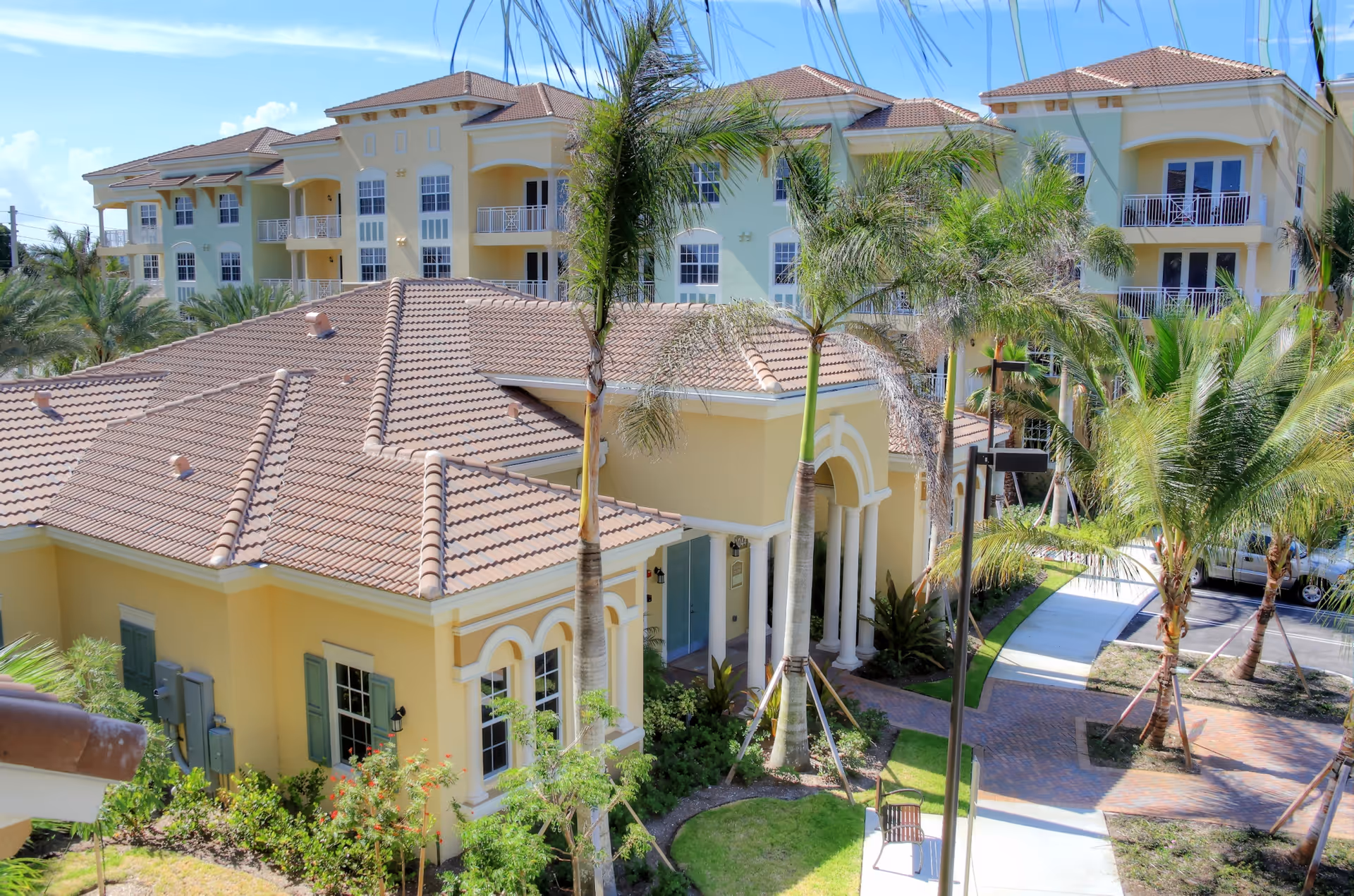 Exterior view of a senior living facility named Riverwalk Pointe featuring a yellow building with a tiled roof, surrounded by palm trees and landscaped greenery. The building has multiple windows and balconies, with a paved walkway and parking area visible.