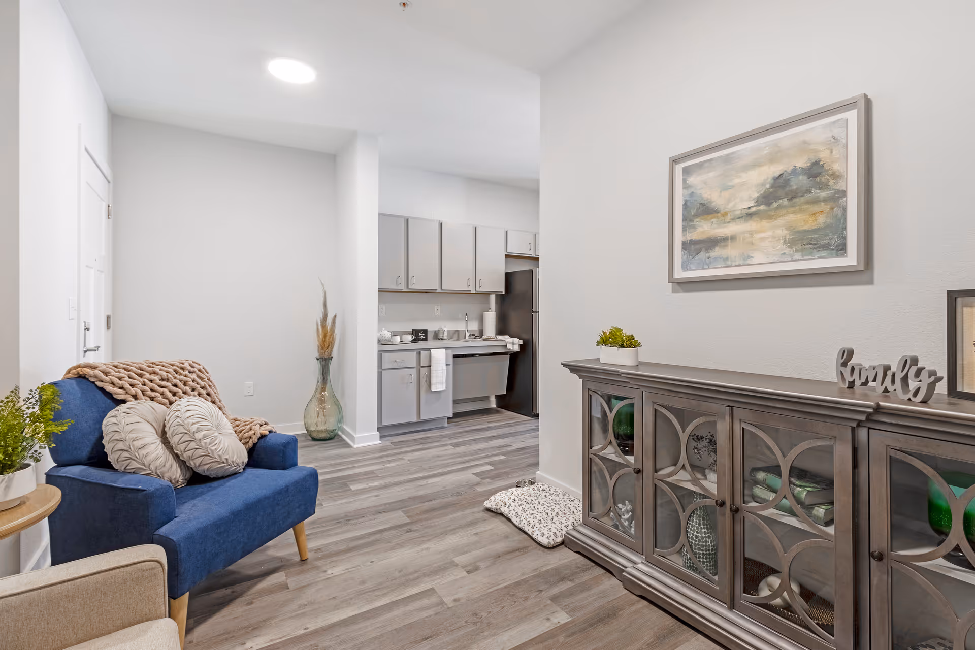 Bright open living area with a blue armchair, decorative cabinet, and a view into a small kitchen.