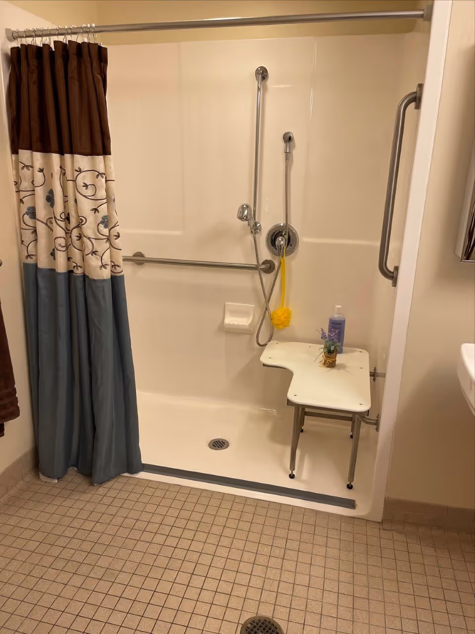 A bathroom shower area with a shower curtain in brown, cream, and blue colors. The shower has a foldable white bench with a small plant and a bottle of body wash on it. There are metal grab bars on the wall for support, a handheld showerhead, and a yellow bath sponge hanging from the shower controls. The floor is tiled with small square tiles in a beige color.