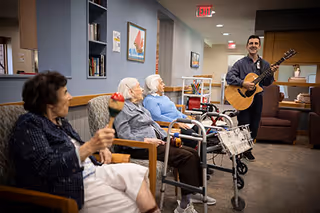 A man playing guitar and singing to three elderly women seated in chairs in a cozy indoor common area. One woman is holding a maraca, and two walkers are visible nearby. The room has bookshelves, framed artwork, and warm lighting.