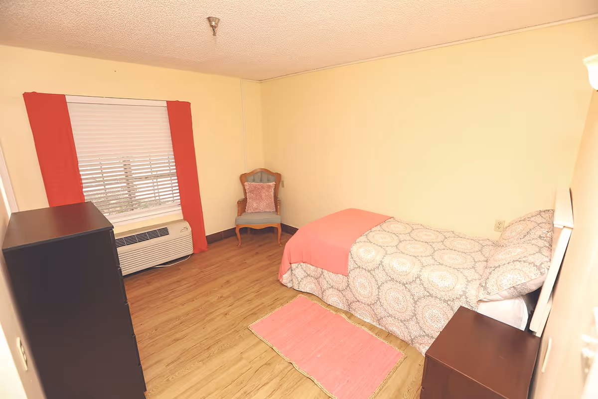 A small bedroom with a single bed covered in patterned bedding and a pink throw blanket. There is a pink rug on the wooden floor beside the bed. A wooden nightstand is next to the bed, and a wooden dresser is positioned near a window with white blinds and red curtains. A cushioned chair with a pink pillow is placed in the corner of the room. The walls are painted light yellow.