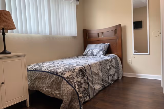 A small bedroom with a single bed featuring a wooden headboard and patterned bedding in shades of gray and white. Next to the bed is a white nightstand with a lamp that has a woven lampshade. A vertical mirror is mounted on the beige wall, and a window with vertical blinds allows natural light into the room. The floor is dark wood.