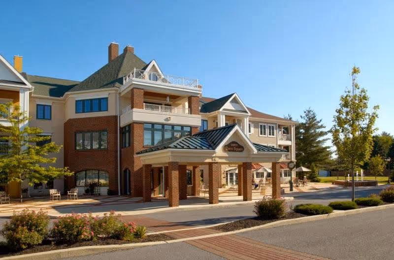 Exterior view of a multi-story senior living facility building with brick and beige siding, a covered entrance with a green roof, landscaped bushes, trees, and a clear blue sky.