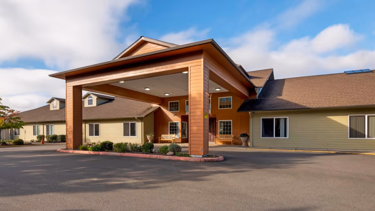 Exterior view of a single-story senior living facility building with a large covered entrance area, beige and brown siding, several windows, and a paved driveway under a partly cloudy sky.