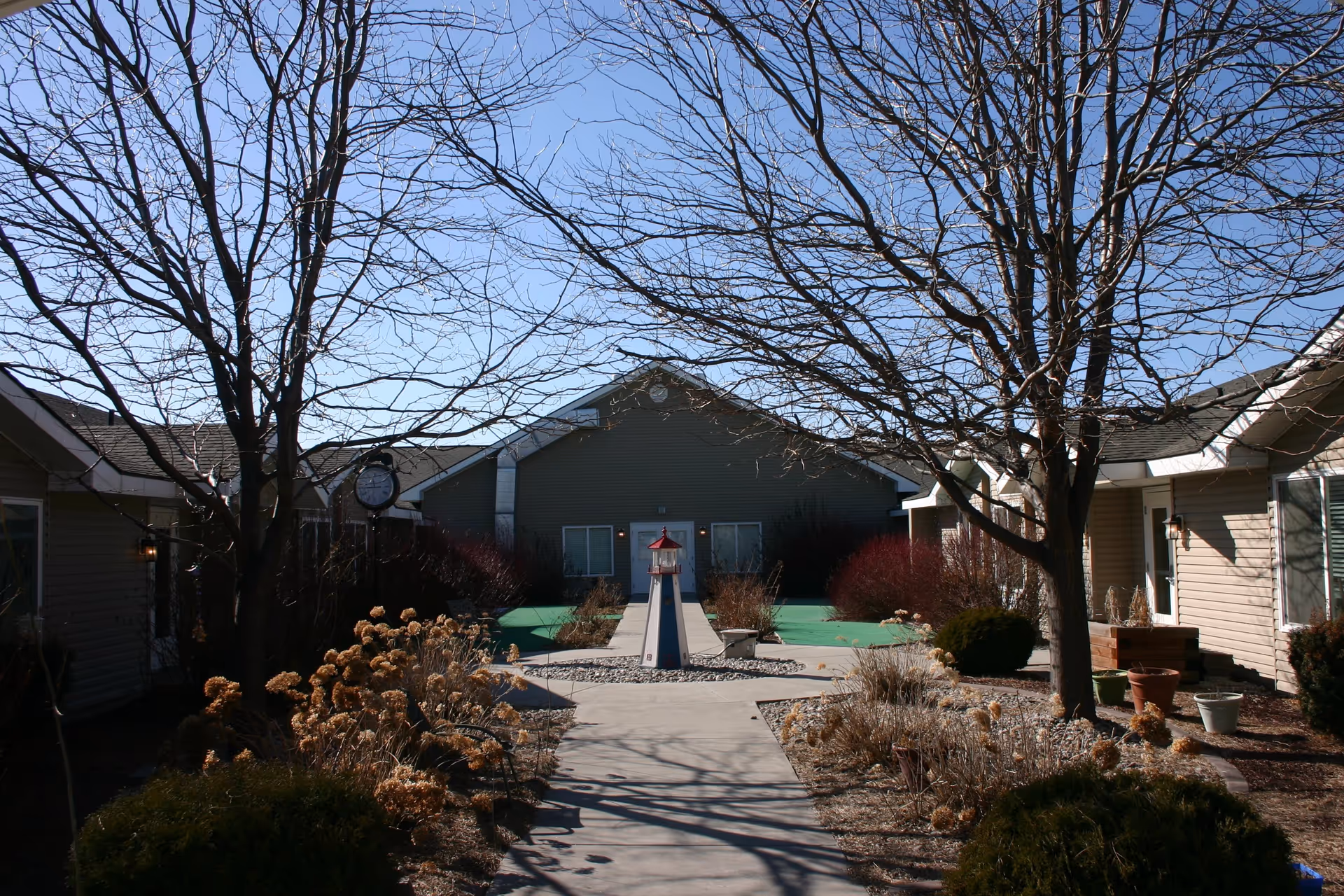 Outdoor courtyard area at Gables of Ammon Assisted Living featuring a paved walkway leading to a small lighthouse decoration in the center. The courtyard is surrounded by leafless trees, dried plants, and single-story buildings under a clear blue sky.