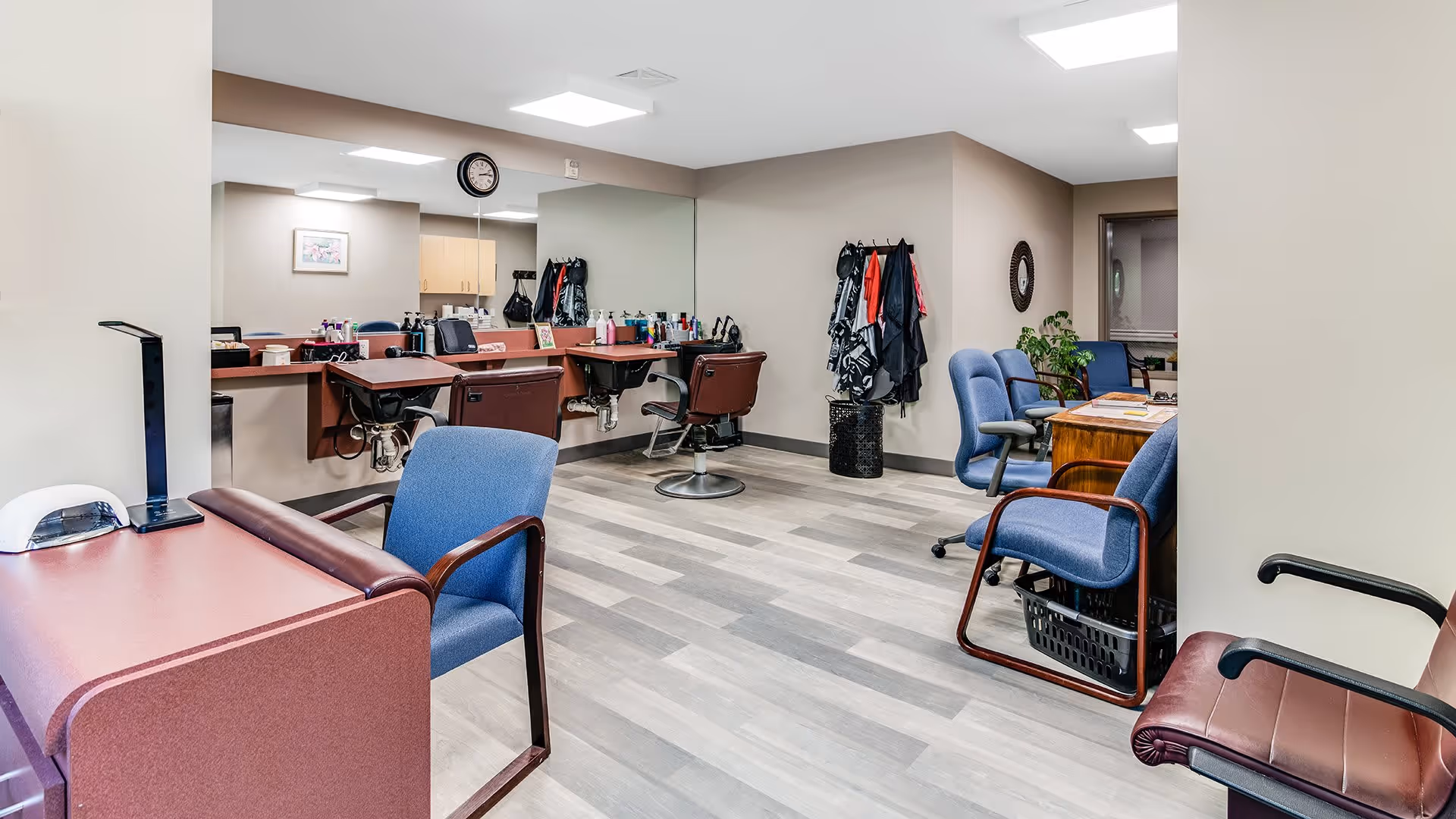 Interior view of a salon area in a senior living facility with salon chairs, mirrors, hair care products on the counter, and several blue and brown chairs arranged around the room.