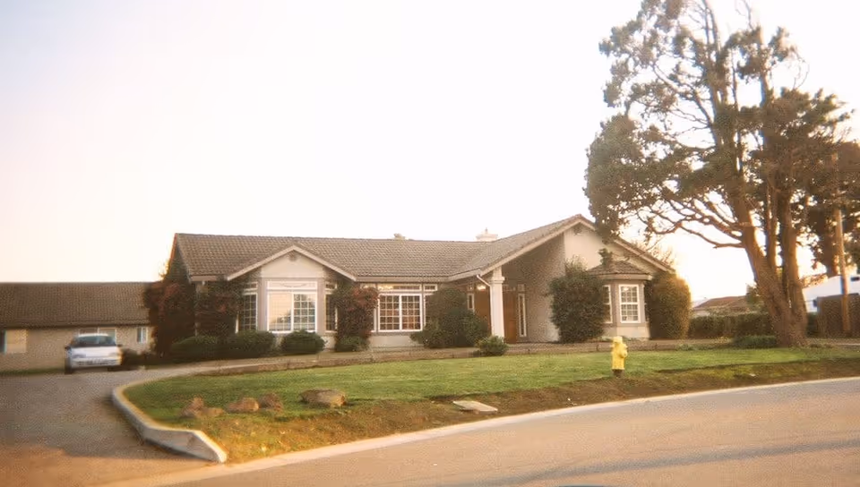 Exterior view of a single-story residential building with a sloped roof, surrounded by bushes and trees, with a driveway and a parked car on the left side and a fire hydrant on the grass near the street.