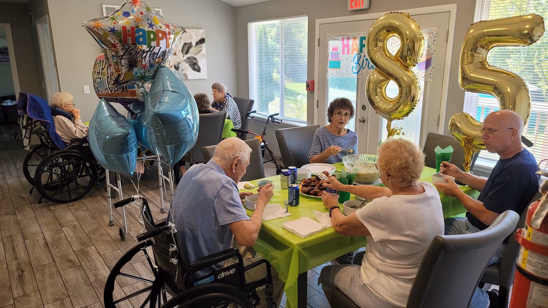 A group of seniors seated around a table eating and celebrating a birthday with balloons, including large gold '85' balloons, in a communal dining area.