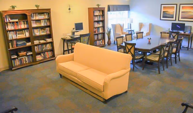 Communal sitting area with a beige sofa, bookshelves, a long table with chairs, and computers in a well-lit room.