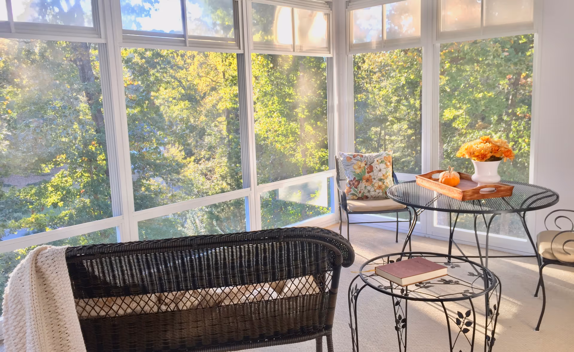 A bright sunroom with large windows showing green trees outside. The room contains a black wicker loveseat with a cream-colored knitted blanket draped over the back, a round black metal table with a wooden tray holding a small pumpkin and a vase of orange flowers, a matching black metal chair with a floral cushion, and a smaller round black metal side table with a closed book on it.