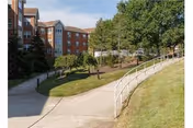 Outdoor view of a senior living facility named Brittany Pointe Estates showing a paved walkway, green grassy areas, trees, and a multi-story brick building in the background under a clear sky.