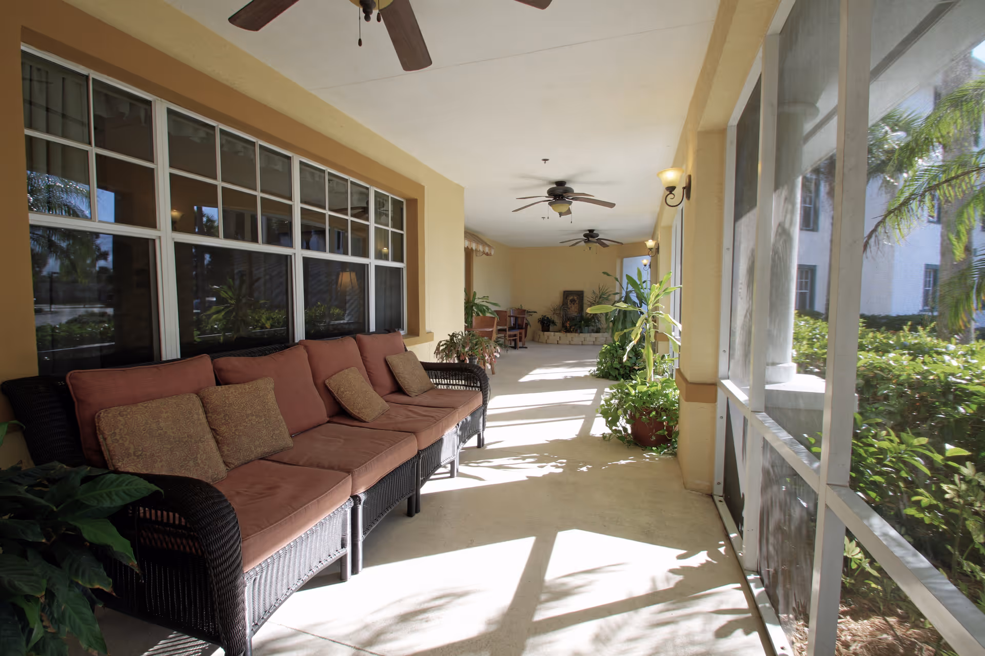 A bright and spacious screened-in porch area with a long cushioned wicker sofa on the left side, several potted plants, ceiling fans, and large windows looking out to greenery and palm trees.