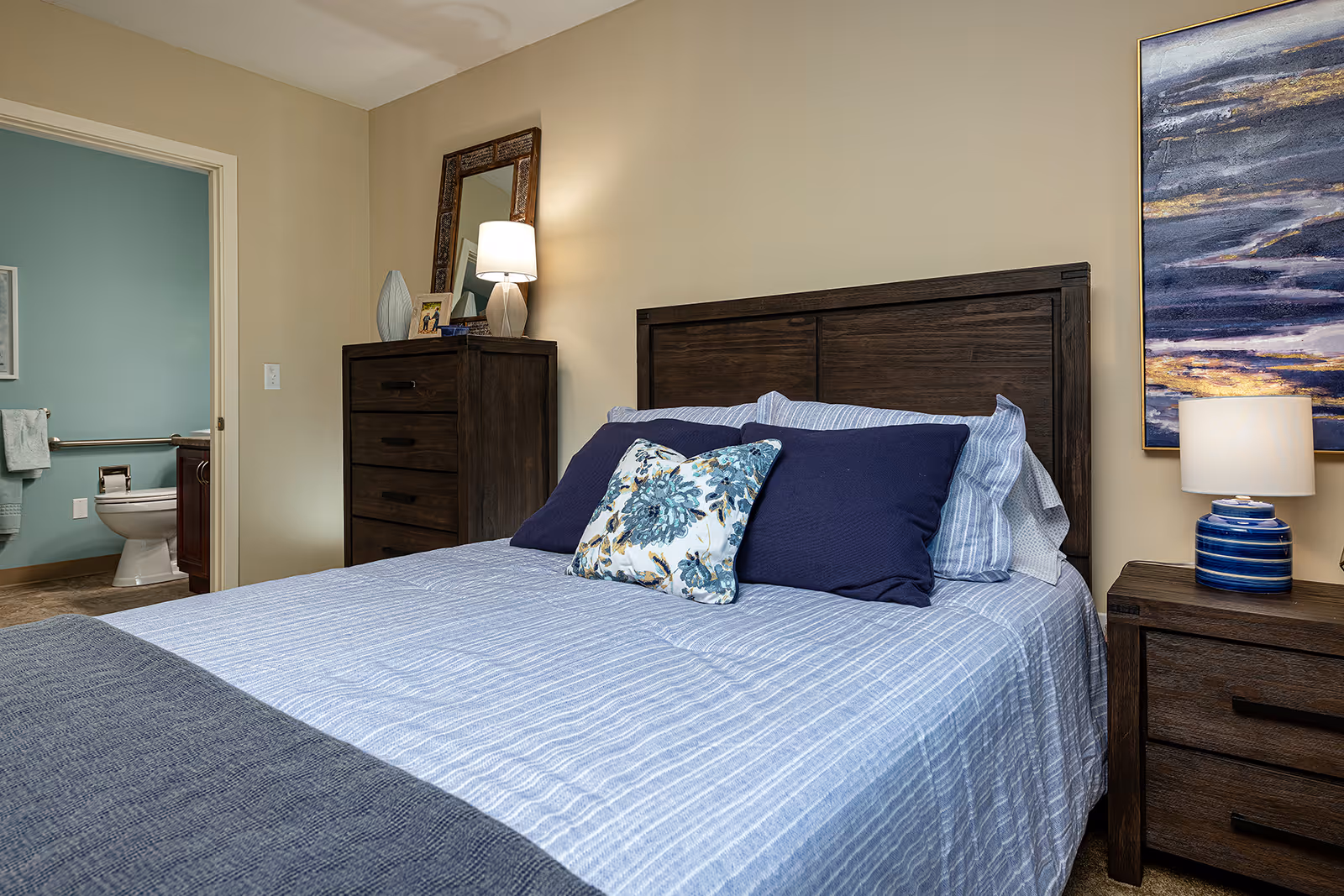 A bedroom with a neatly made bed featuring blue and white striped bedding and decorative pillows. To the right of the bed is a wooden nightstand with a blue and white lamp and a large abstract painting on the wall above it. On the left side of the room is a wooden dresser with a lamp, a mirror, and decorative items on top. An open door reveals a bathroom with a toilet, towel rack, and light blue walls.