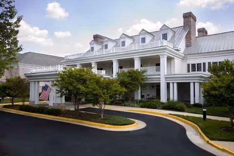White, multi-story senior living facility with wraparound porch, curved driveway, and manicured landscaping.