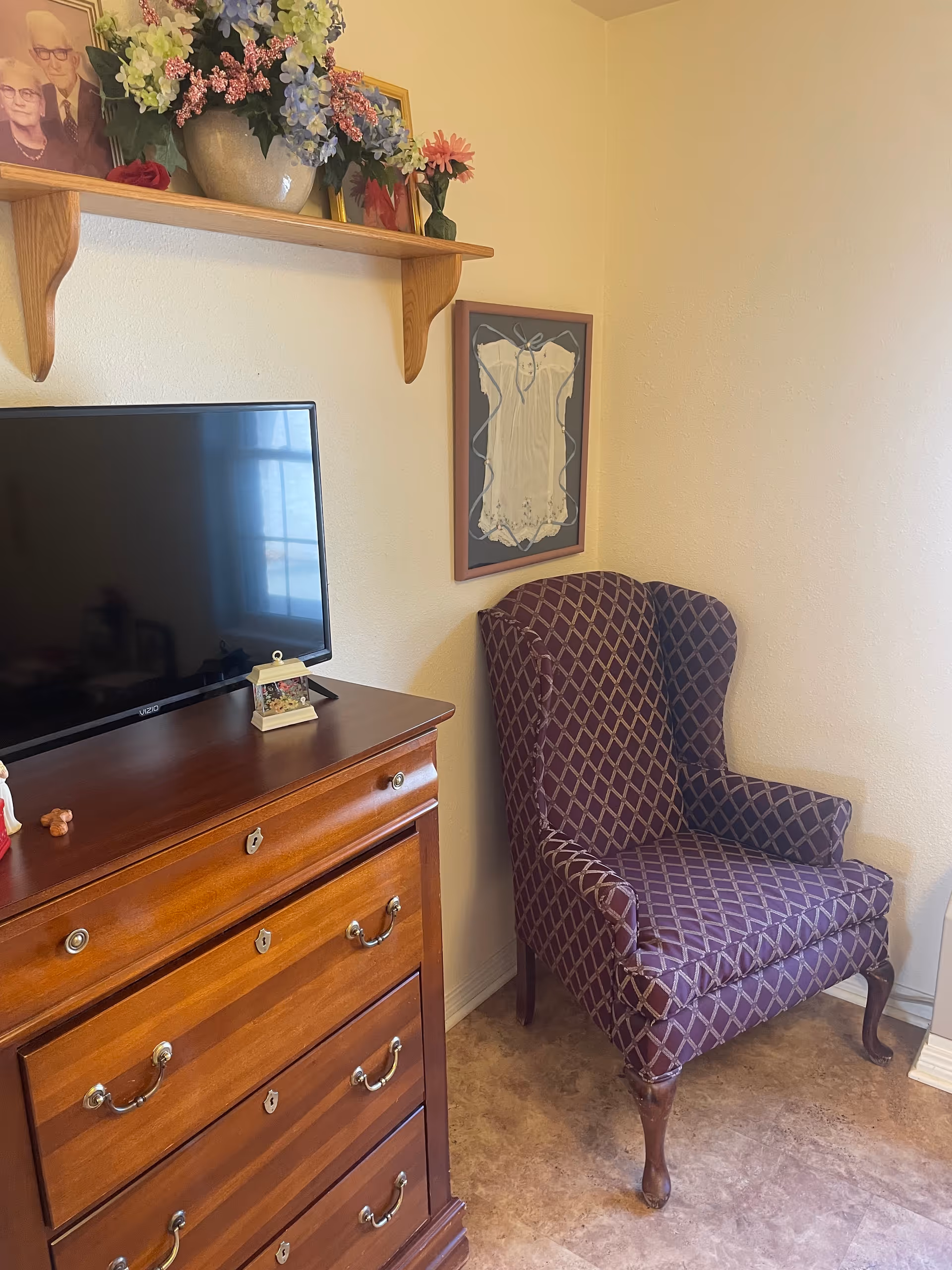 A corner of a room featuring a wooden dresser with a flat-screen TV on top, a purple patterned wingback chair, and a wooden wall shelf holding a vase with colorful flowers and framed photographs. A framed white garment is displayed on the wall above the chair.