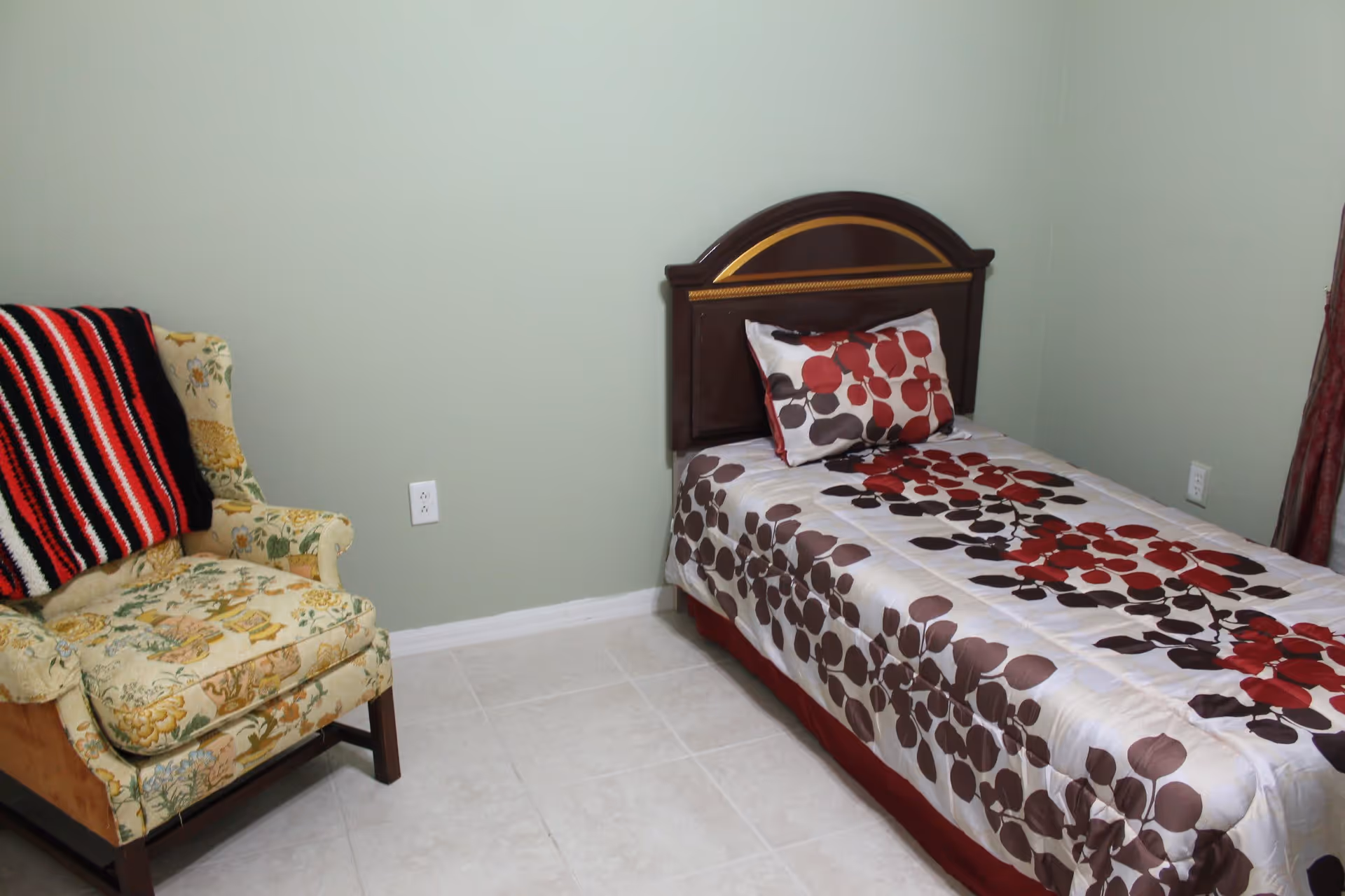 A small bedroom with a single bed featuring a floral patterned bedspread in red, brown, and white colors. Next to the bed is a floral upholstered armchair with a red, black, and white striped blanket draped over it. The walls are painted light green and the floor is tiled in a light color.