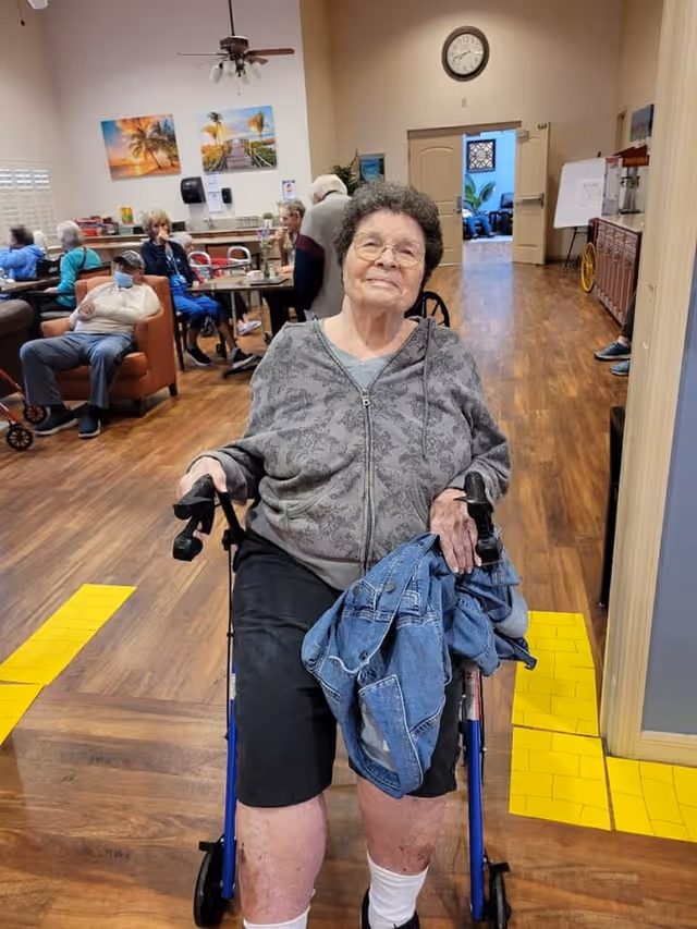 An elderly woman sitting on a blue walker in a senior living facility common area. She is wearing glasses, a gray patterned zip-up jacket, black shorts, and white socks, holding a denim jacket on her lap. In the background, several other elderly people are seated on chairs and sofas, with wall art, a clock, and a ceiling fan visible in the room.