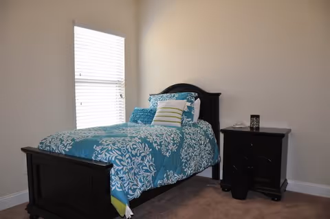 A small bedroom with a single bed featuring a dark wooden frame and blue floral bedding. There are multiple pillows on the bed, including a white pillow with green stripes. Next to the bed is a dark wooden nightstand with a white telephone and a decorative item on top. The room has beige walls, a window with white blinds, and carpeted flooring.