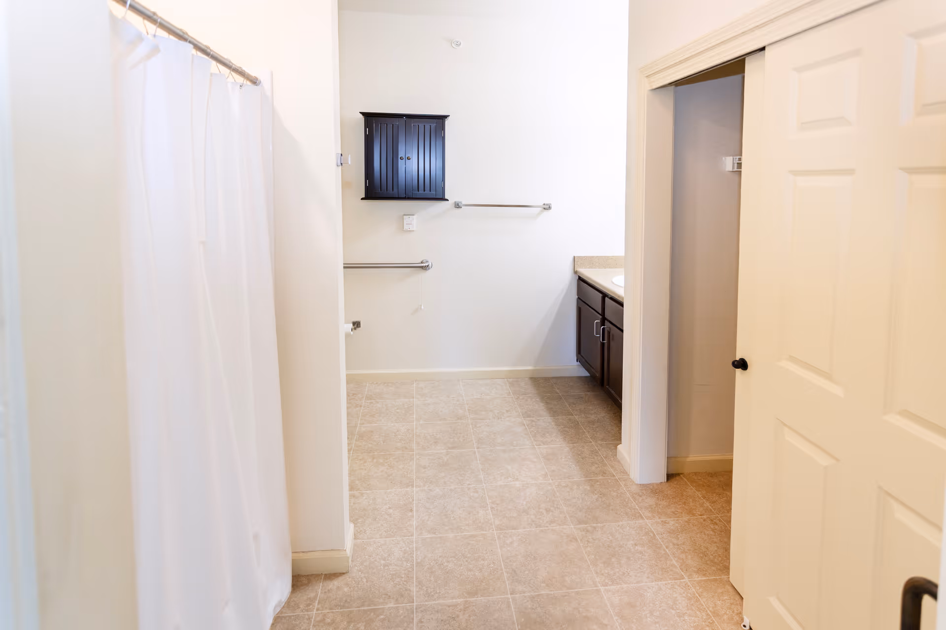 A clean bathroom with beige tiled floor, a white shower curtain on the left, a dark wooden cabinet mounted on the wall, towel bars, and a countertop with dark cabinets underneath on the right. There is also a partially open door leading to a closet or another room.