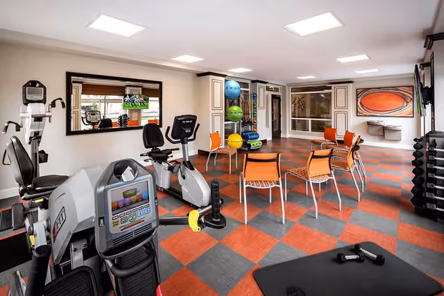 A bright fitness room with exercise equipment including stationary bikes, weights, and a rack of colorful exercise balls. The room has a checkered red and gray floor, several orange chairs arranged in a semi-circle, large windows, and a wall-mounted TV displaying a sports game.