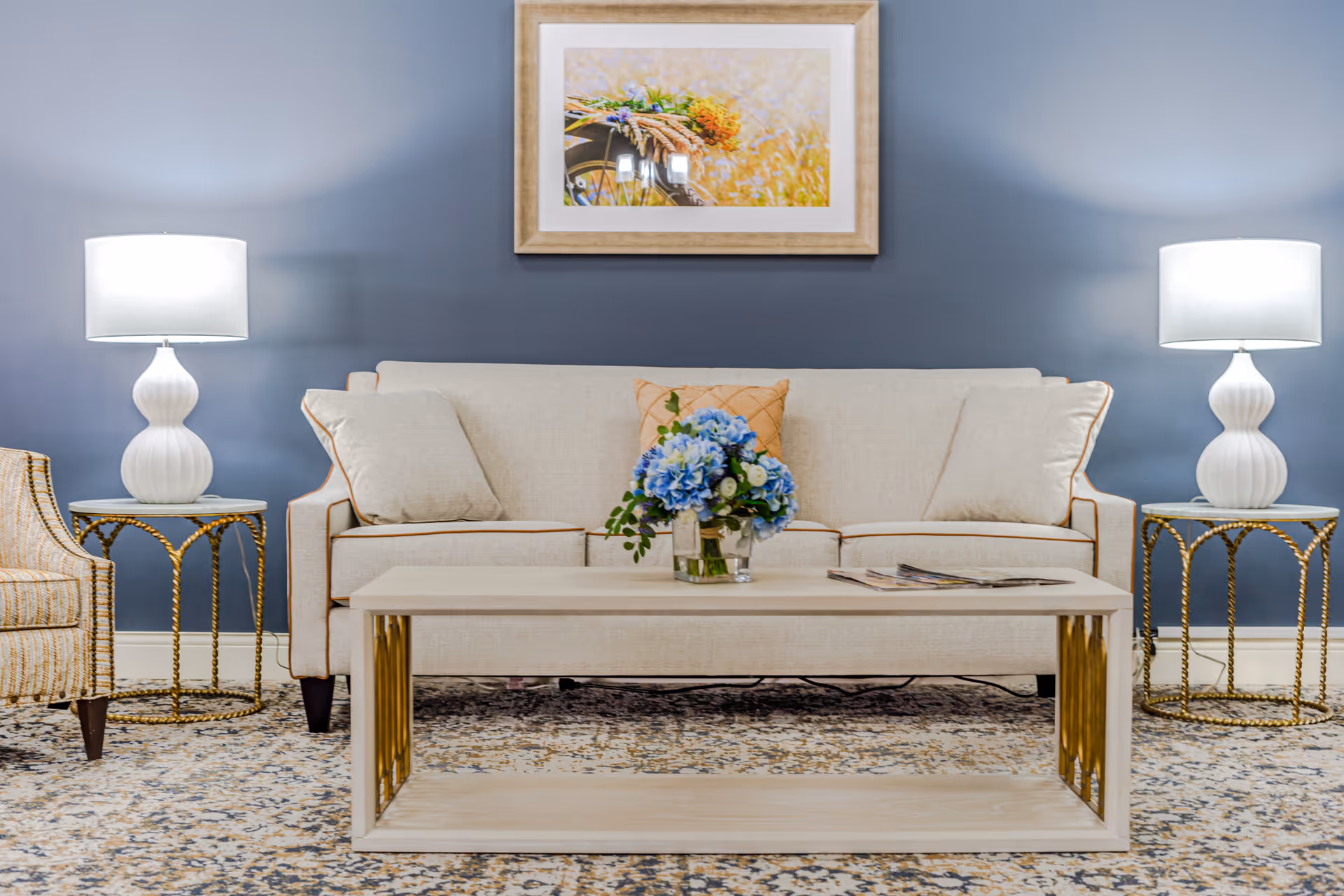 A cozy living room with a beige sofa adorned with three cushions, a light wooden coffee table with a vase of blue and white flowers, two white table lamps on gold-accented side tables, a patterned carpet, and a framed picture of flowers on a blue wall.