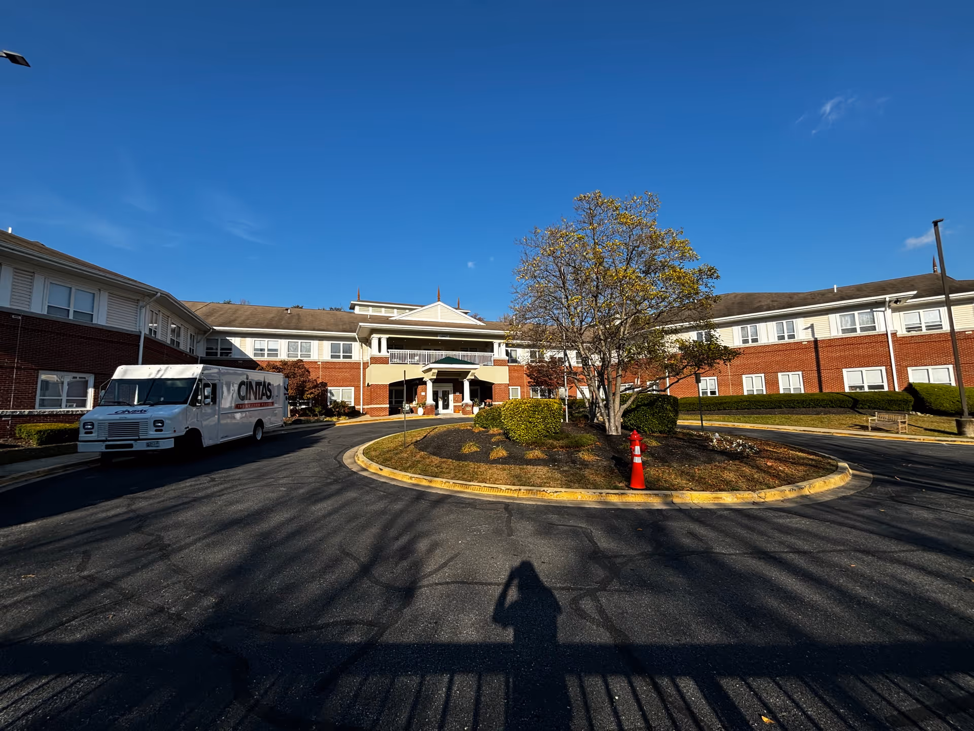 Front exterior view of a two-story senior living facility building with red brick and beige siding. There is a circular driveway with a landscaped island in the center featuring a tree and bushes. A white delivery truck is parked on the left side of the driveway. The sky is clear and blue.