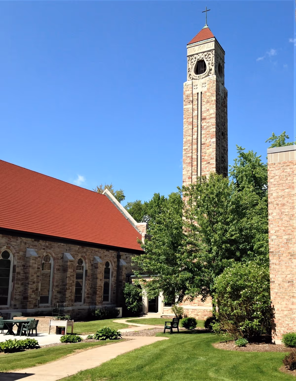 Outdoor view of a building with a tall stone tower featuring a cross on top and the words 'SING JOYFULLY' near the top. The building has a red sloped roof and arched windows. There is a well-maintained lawn with a paved walkway, green bushes, trees, and outdoor seating including a table with chairs and a bench.