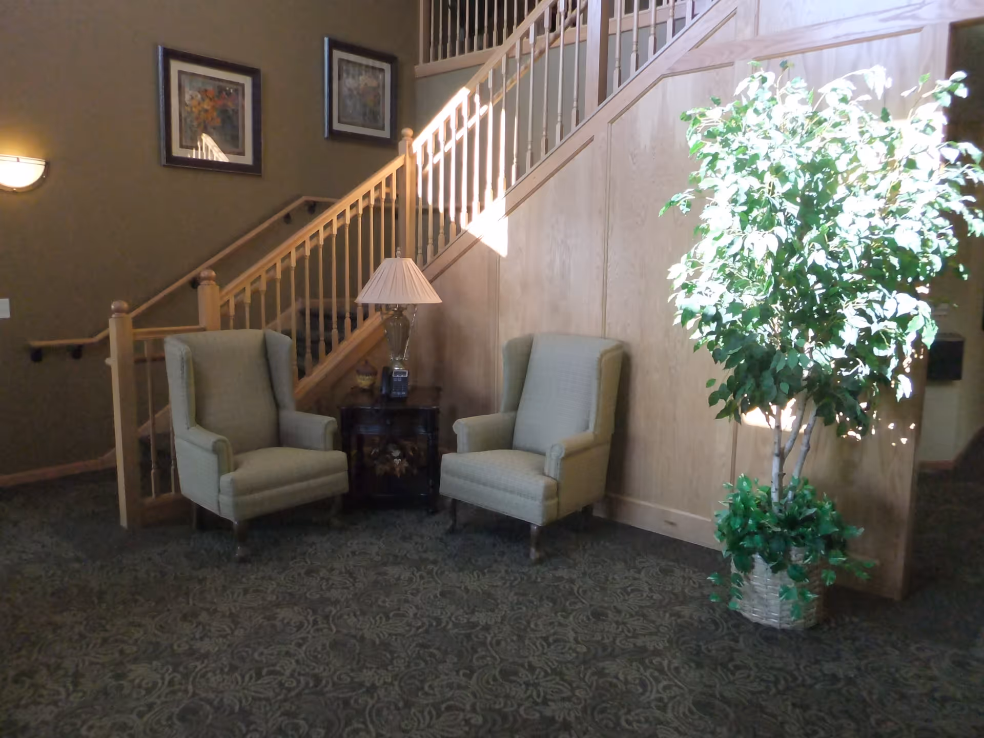 Two upholstered armchairs and a side table with a lamp beside a staircase and a large potted plant in a lobby.